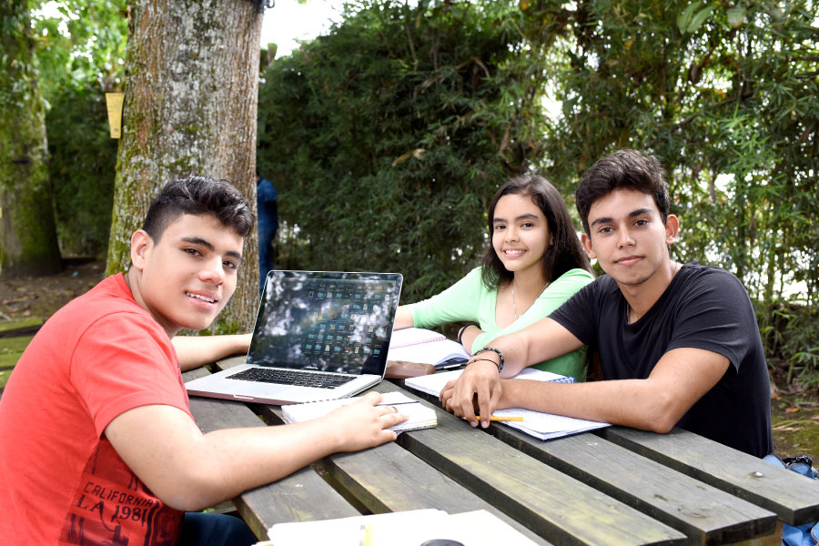 Estudiantes de universidad sonriendo a la cámara y sentados estudiando con libros, cuadernos y portátil