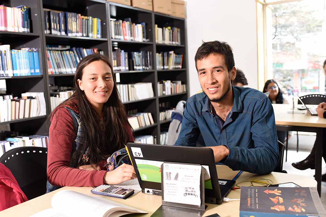 Imagen de dos estudiantes sentados en la biblioteca sonriendo mientras usan el computador