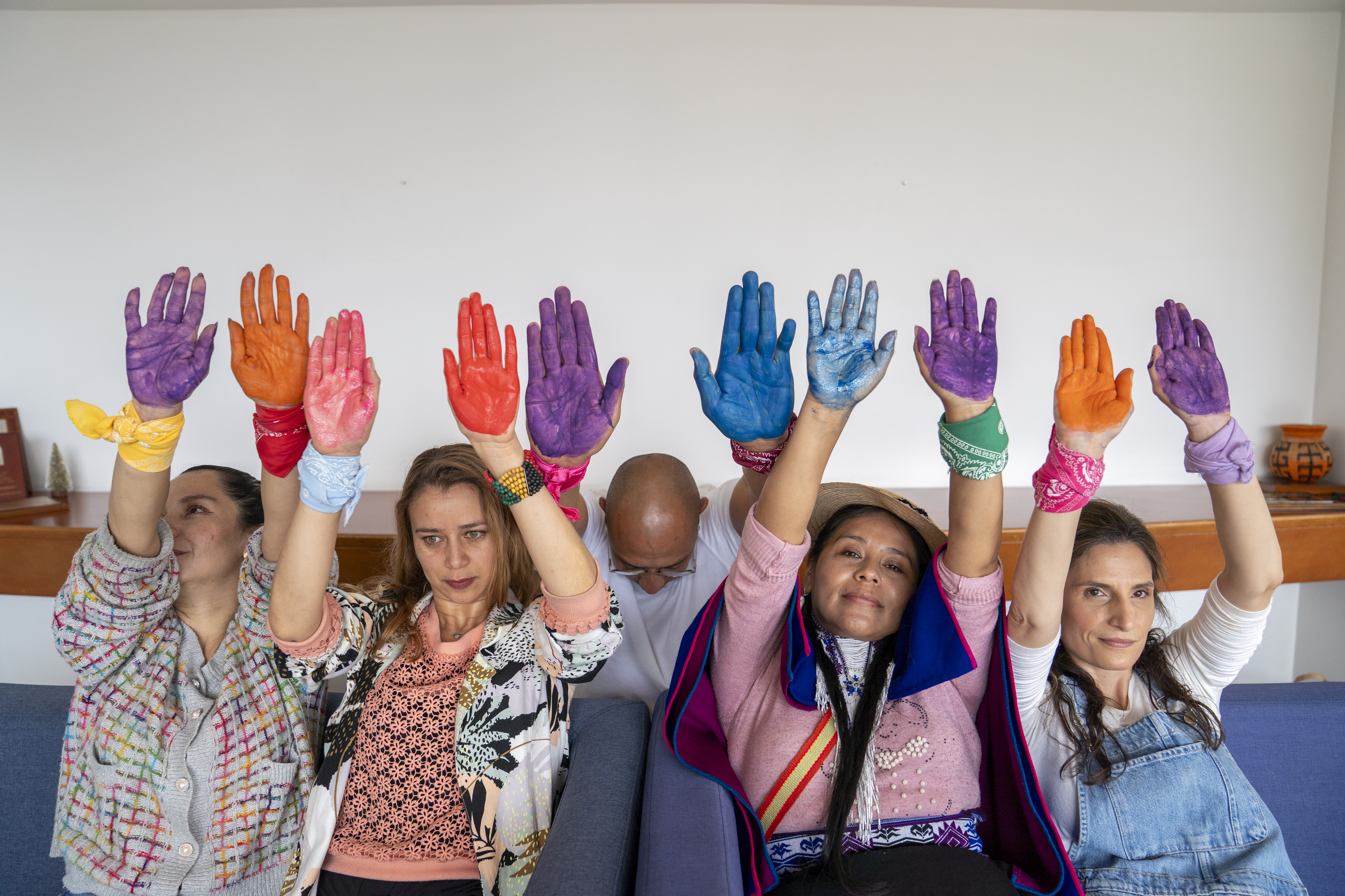 5 personas, un hombre y cuatro mujeres de diferentes etnias y con las manos arriba con las palmas hacia el frente, pañoletas en las muñecas, y las palmas con pintura del arcoíris